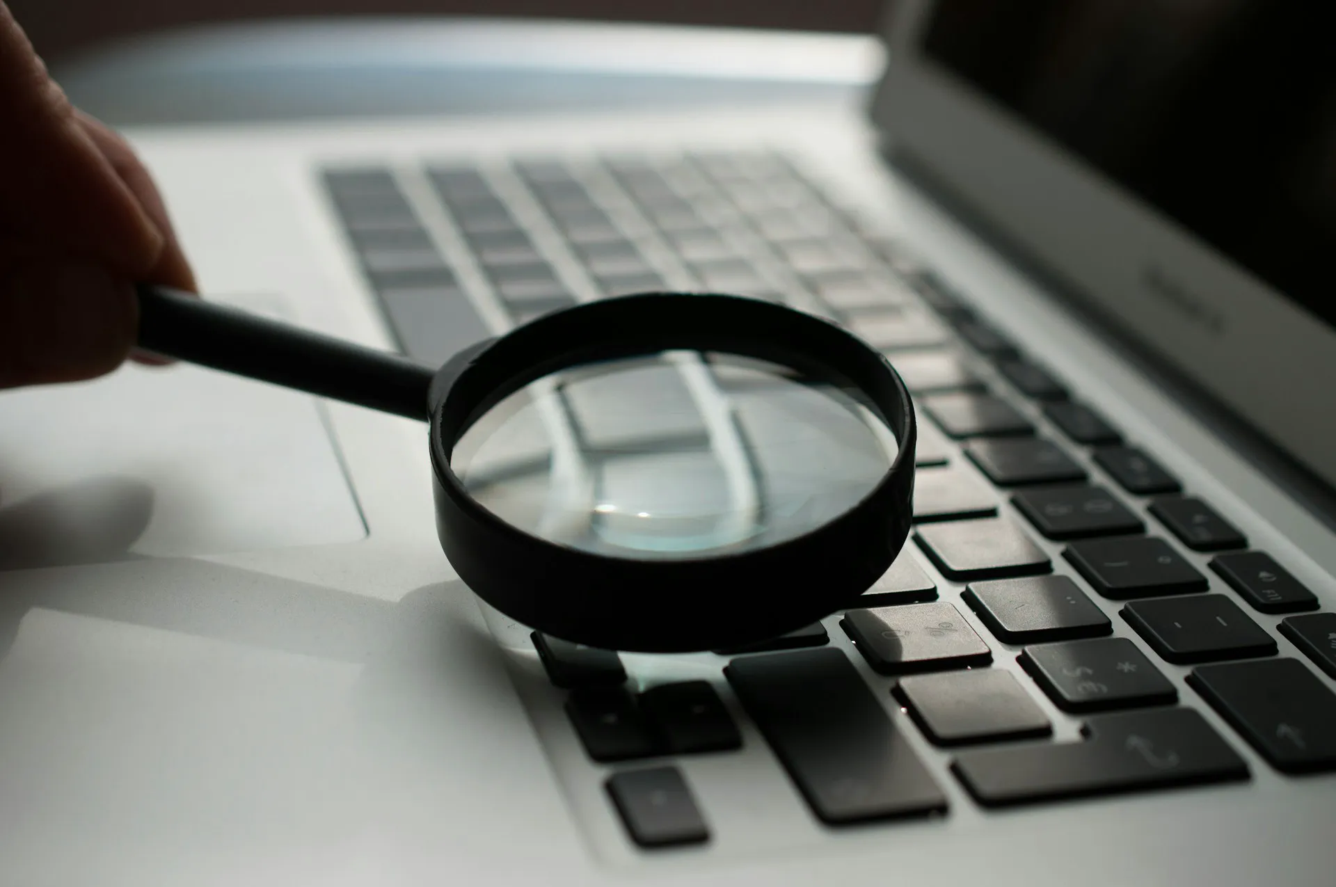 A hand holding a magnifying glass over a laptop keyboard, illustrating attention to detail and thorough inspections